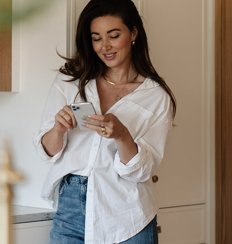 Woman in a white shirt and blue jeans using a smartphone in a kitchen.