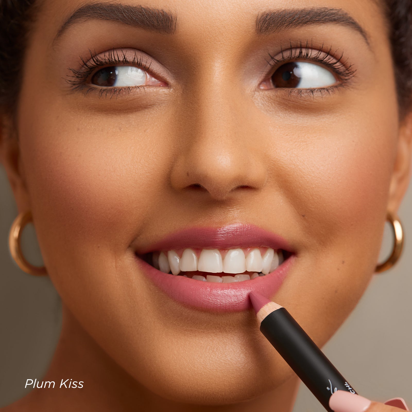 Woman applying plum lip color with a pencil, wearing gold hoop earrings on a neutral background.