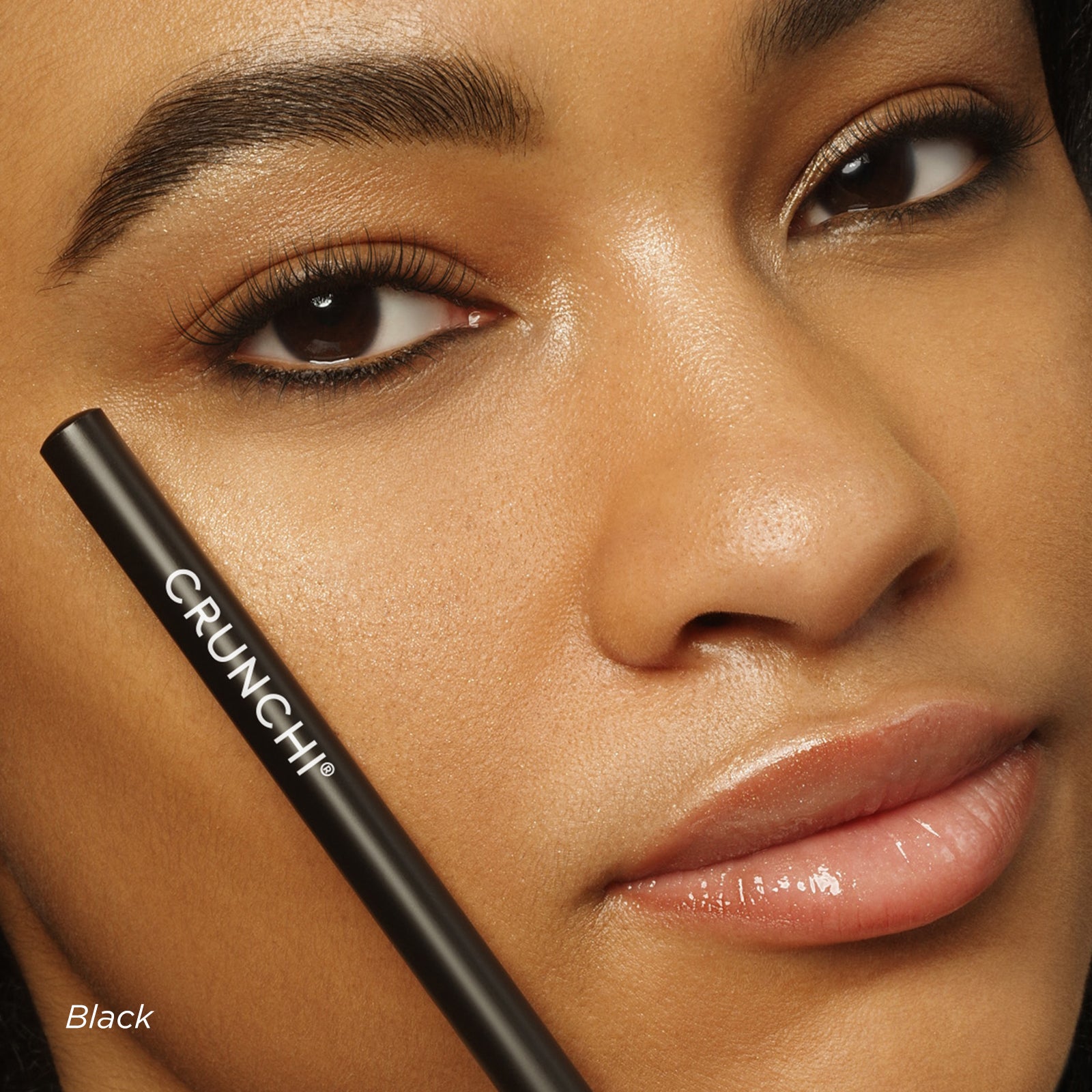 Close-up of a woman's face with a black eyeliner pencil labeled 'CRUNCHI' on a neutral background.